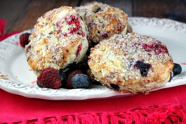 three berry scones on a white platter with fresh berries scattered. Pink cloth napkin underneath