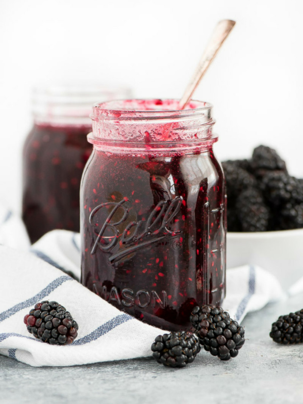 jar of blackberry freezer jam with a spoon in it. white napkin with blue plaid underneath. fresh blackberries scattered around