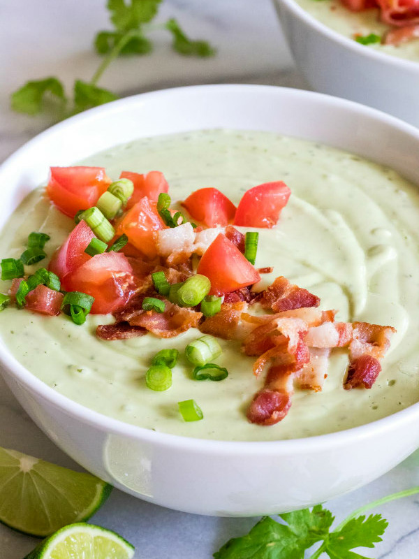 chilled avocado soup in a white bowl topped with green onion and tomato. lime wedges, cilantro and another bowl of soup in the background