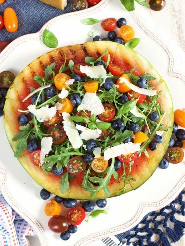 Grilled Watermelon Pizza with Blueberries, Parmesan and Arugula on a white plate with more tomatoes and fruit and a wedge of parmesan in the background