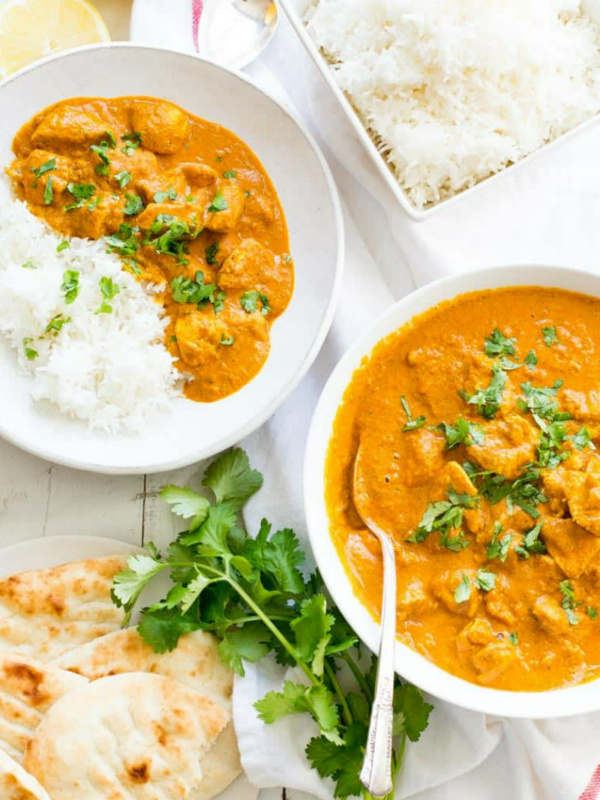 overhead shot of two servings of indian butter chicken in white bowls with white rice and cilantro garnish and pita bread on the side