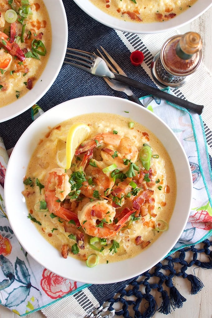 overhead shot of shrimp and grits in a white bowl on a patterned napkin with a fork and two other partial views of shrimp and grits bowls