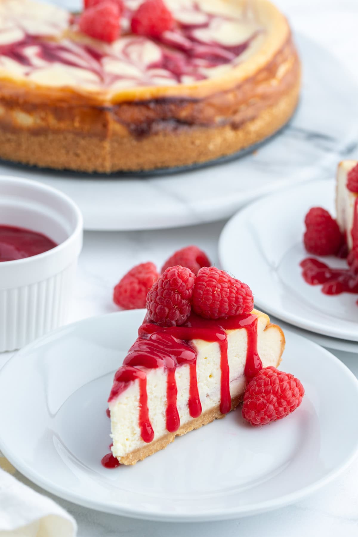 slice of white chocolate raspberry cheesecake on plate with whole cheesecake in background