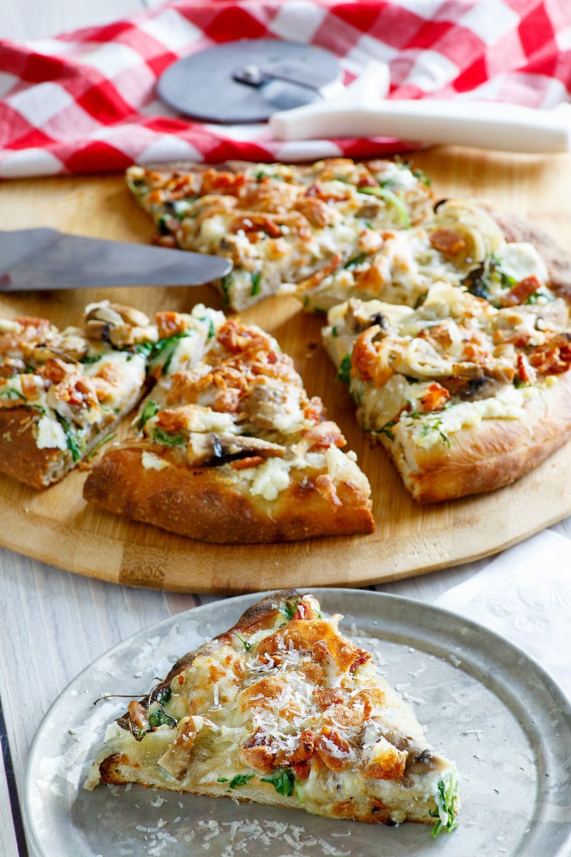 slice of white pizza on a metal plate with cut pizza on a cutting board in the background with red and white checked napkin and pizza cutter